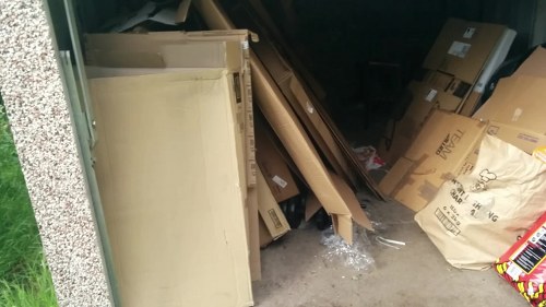 A cluttered indoor or garage space with a collection of discarded wooden furniture, cardboard boxes, and packaging materials. On the left side of the image, there is a partially visible cream-colored wooden cabinet or shelving unit leaning against a wall with a textured exterior wall nearby. In the center and right portions of the space, various items such as flattened cardboard boxes, some with printed logos, and crumpled packing paper are stacked haphazardly, occupying the floor area. The items appear to be sorted for disposal or removal, with some boxes showing signs of wear or damage. The environment suggests an area used for storing waste materials awaiting collection, consistent with rubbish removal services from Business Waste Removal Haringey. The scene is illuminated by natural or ambient lighting, highlighting the textures of the cardboard and wood, with the background obscured by darker elements and additional boxes. This setup aligns with typical rubbish clearance tasks in residential or small commercial settings in North London, nearby Haringey.