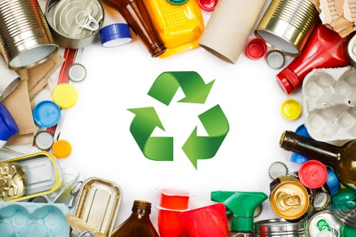 A variety of discarded recyclable waste materials arranged in a circular pattern around a prominent green recycling symbol in the center. The waste includes aluminum cans with metallic finishes in silver, gold, and red, plastic bottles in blue, yellow, green, and brown, some with caps attached, as well as plastic containers and packaging in red, green, and other colours. Cardboard tubes and flattened cardboard pieces are visible, along with some crumpled plastic bags. The background is plain white, emphasizing the clean, organized appearance of the waste items. The scene suggests responsible waste sorting and recycling practices, relevant to rubbish removal services in Haringey, London, and the surrounding areas. Business Waste Removal Haringey specializes in managing and disposing of such waste materials in compliance with local recycling standards, aiding businesses in sustainable waste management efforts.