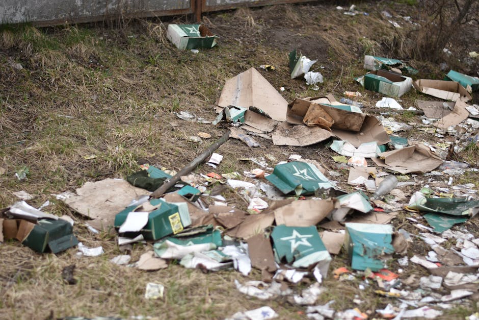 A roadside scene showing a pile of mixed waste and rubbish, including black plastic bags, a yellow plastic container, and a discarded sofa cushion with fabric upholstery, all resting on a gravel surfa