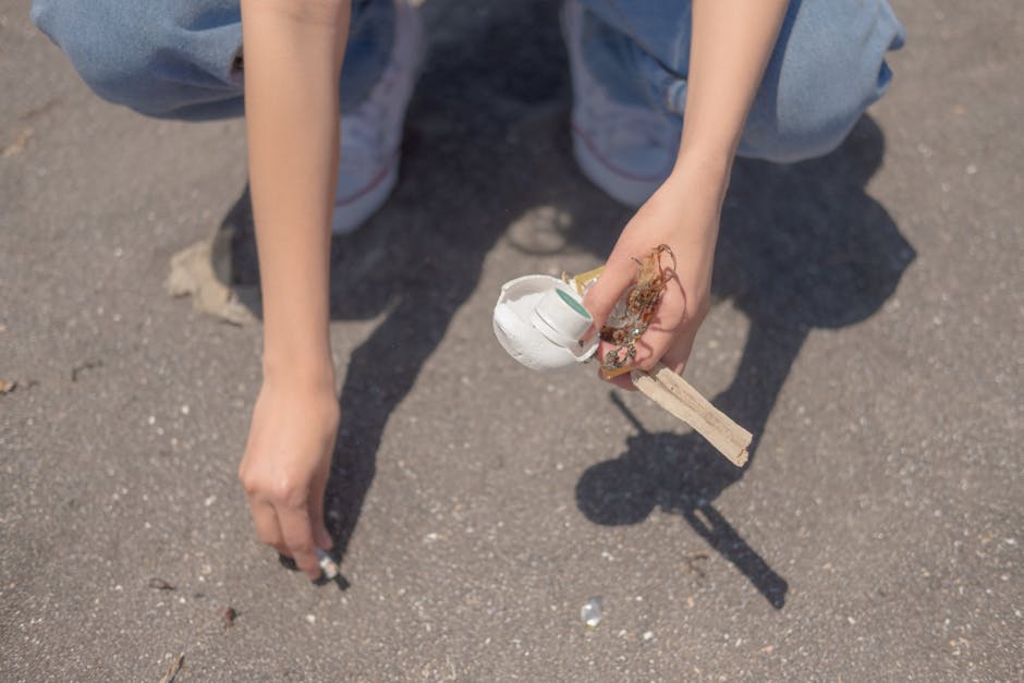 A close-up view of a person's hands crouching on a paved outdoor surface, with one hand reaching towards the ground and the other holding a small wooden stick with a crumpled white piece of paper and 